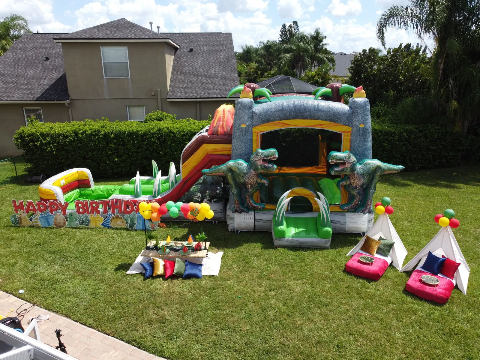 Dinosaur birthday party setup with a dino bounce combo, picnic tables and teepees on a backyard lawn